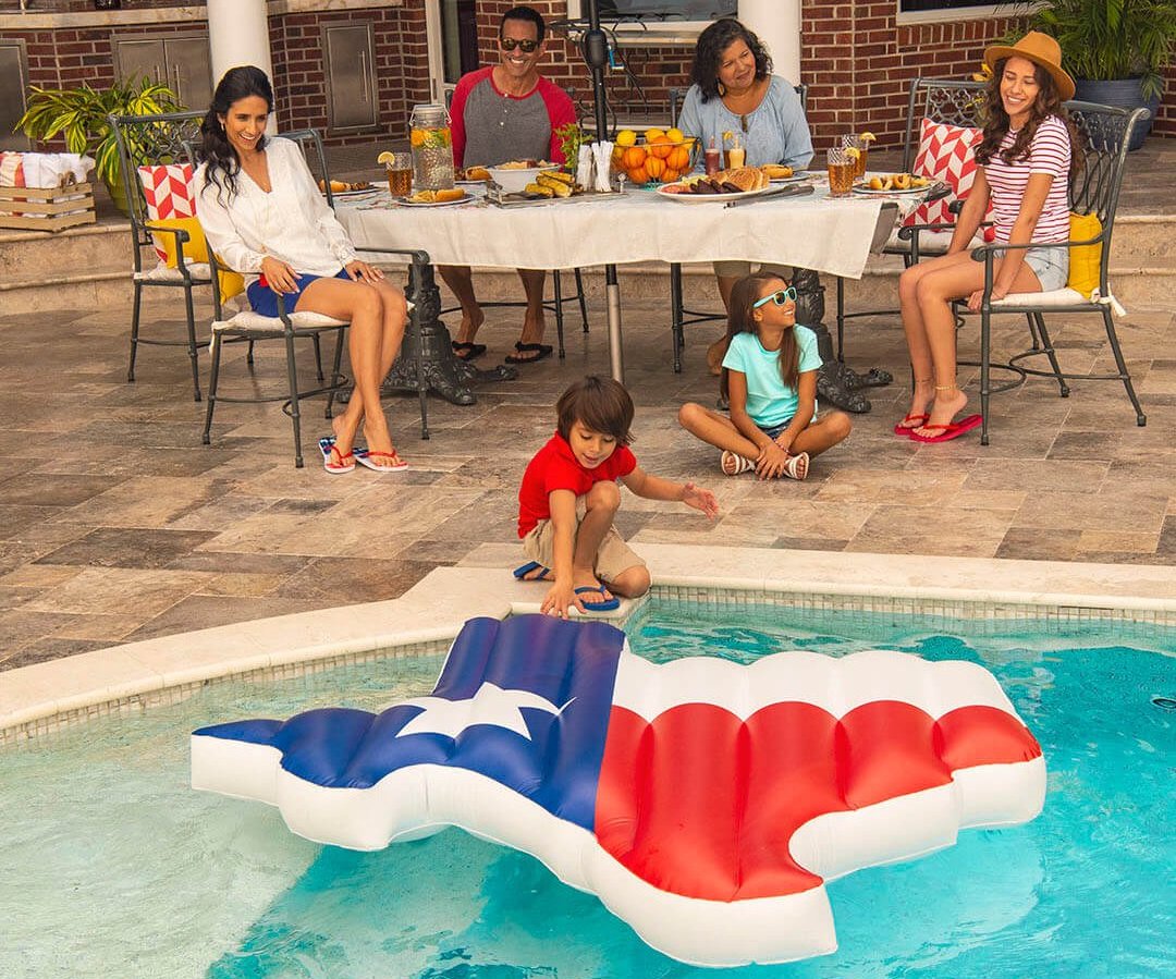Family enjoying a meal on the patio beside a swimming pool with a large Texas-shaped pool float in red, white, and blue colors resembling the state flag.