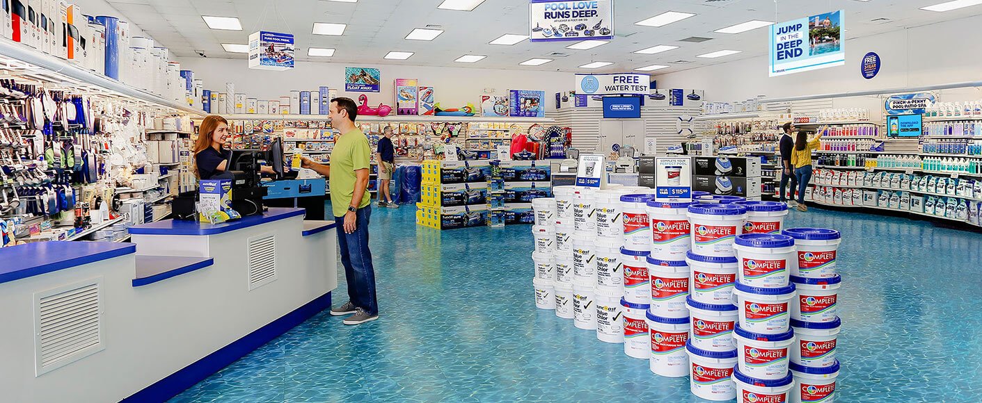 Customer shopping inside a brightly lit Pinch A Penny store with stacks of pool chemical buckets displayed near the checkout counter and aisles filled with pool supplies.