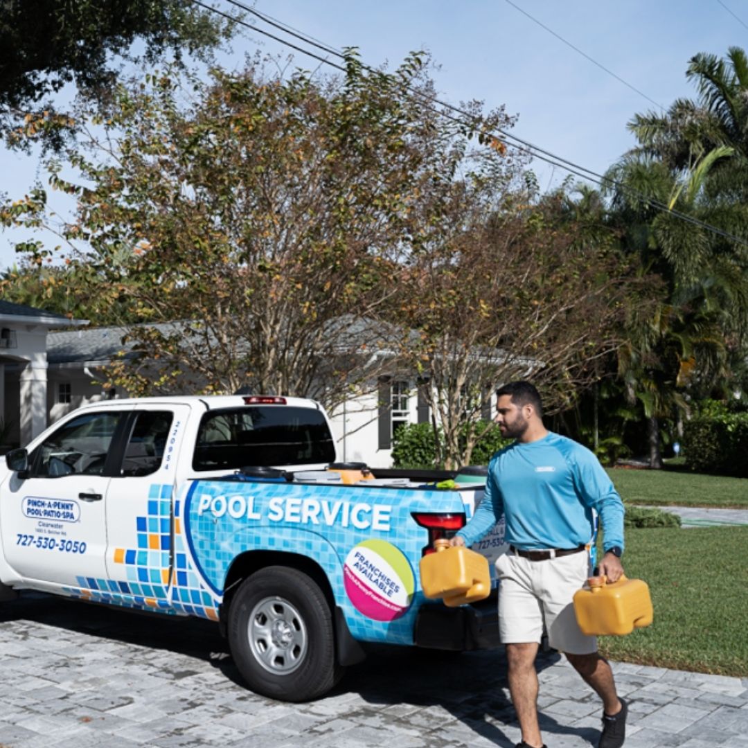Pinch A Penny pool service technician walks beside a branded service truck carrying two yellow chemical jugs.