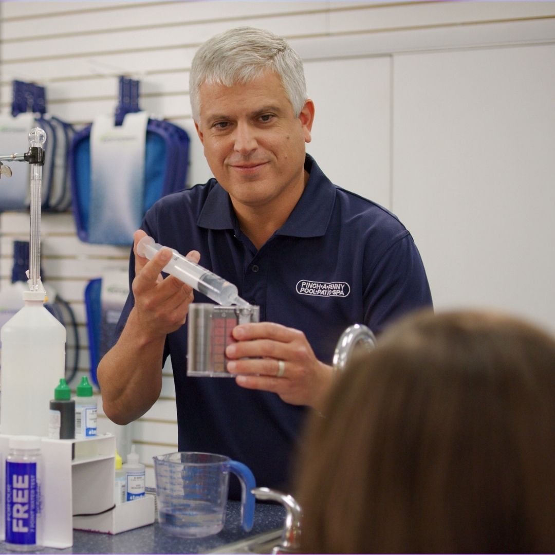 Pinch A Penny employee demonstrates a pool water test, holding a syringe and color comparator block at a counter while a customer watches.
