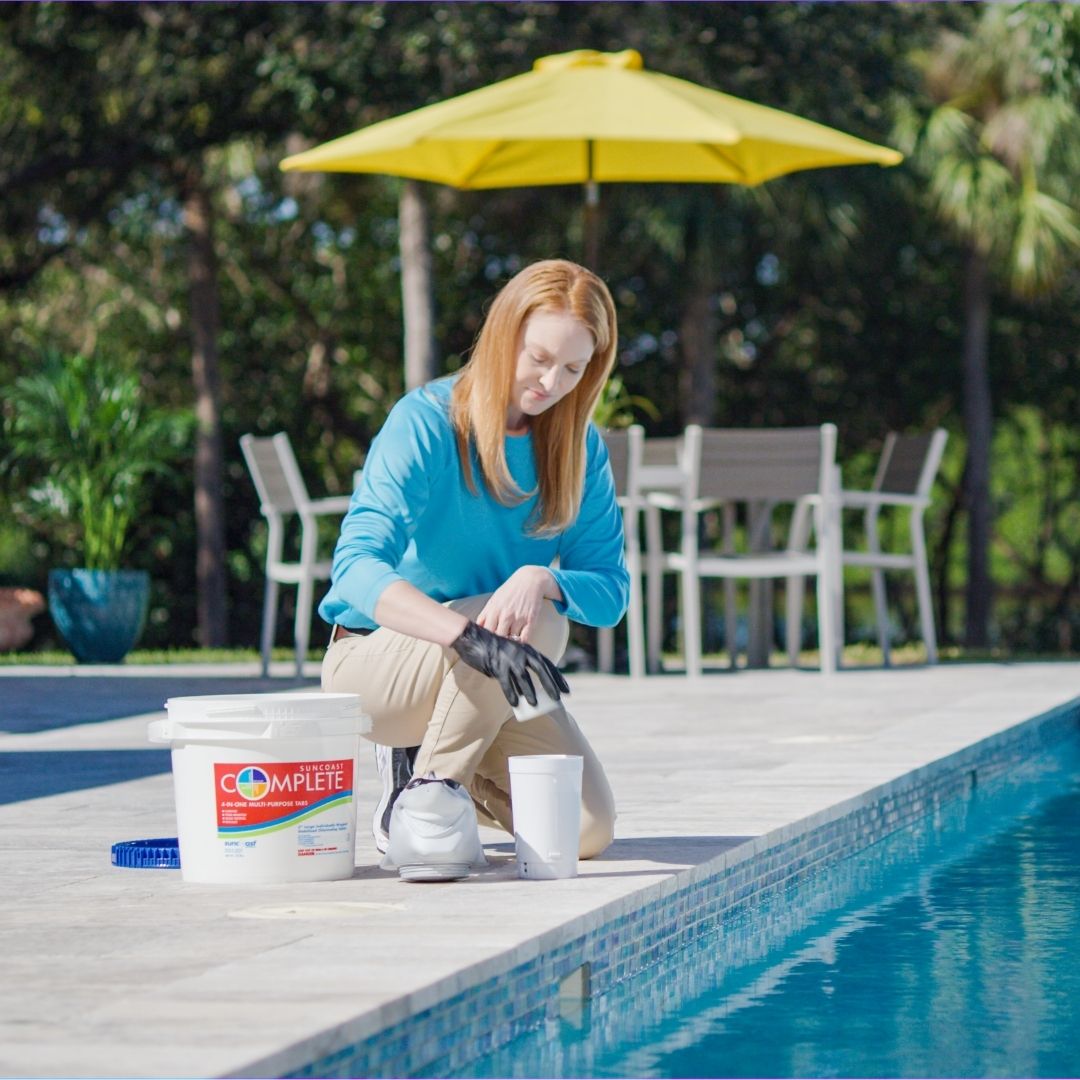 Pinch A Penny technician kneels at the edge of a pool measuring or dispensing pool chemicals from a bucket during routine maintenance.