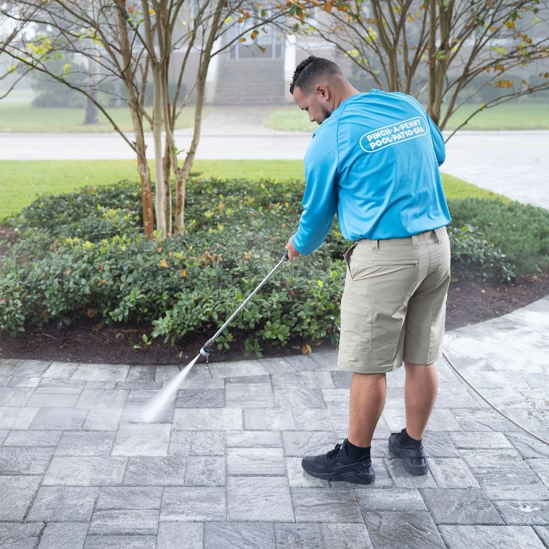 Pinch A Penny technician in uniform pressure-washes a paved driveway or patio with a spray wand.