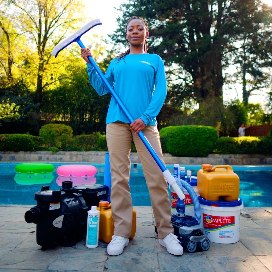 Pinch A Penny technician stands poolside holding a pool brush, with pool equipment and maintenance products arranged nearby.