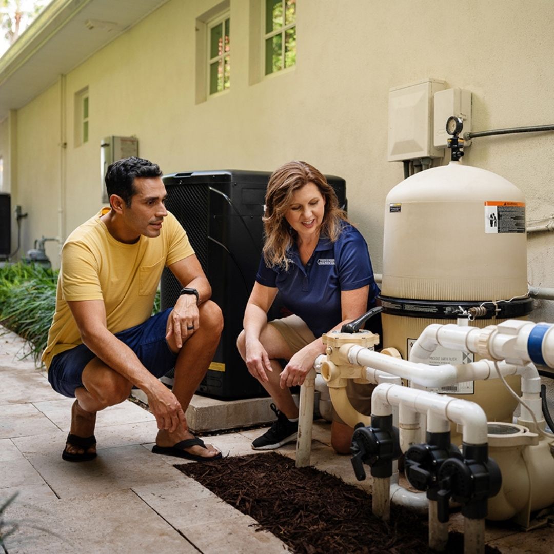 Two people crouch beside pool equipment, looking at valves and plumbing on a filter system during a pool equipment check.