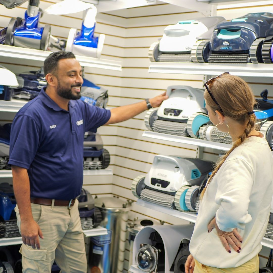 Pinch A Penny store associate points out robotic pool cleaner models on a display wall while assisting a shopper.