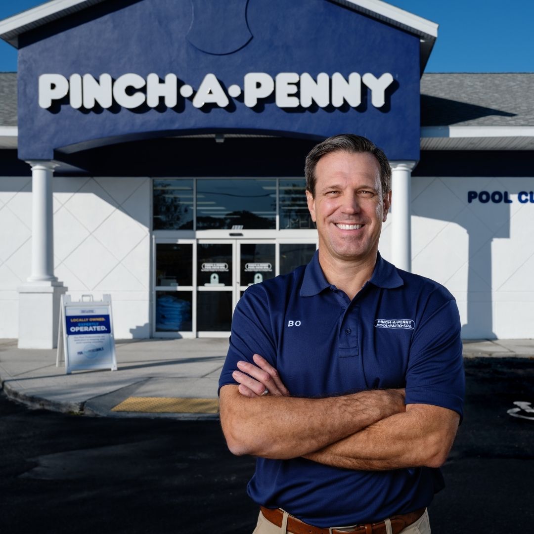 Pinch A Penny franchise owner stands with arms crossed in front of the store entrance and large Pinch A Penny sign.