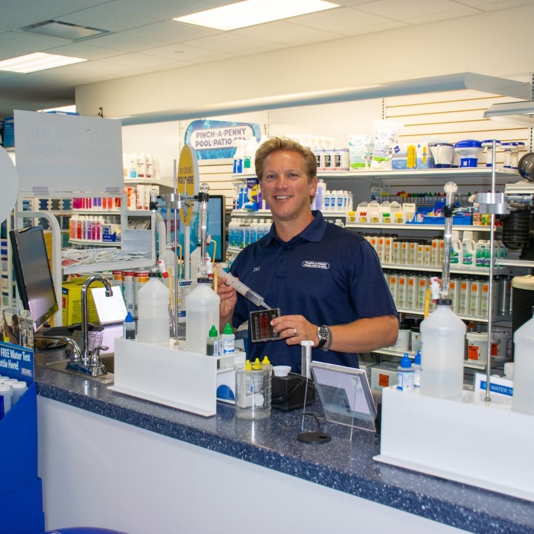 Pinch A Penny employee performs a pool water test at a counter, holding a syringe and test block while explaining results to a customer.