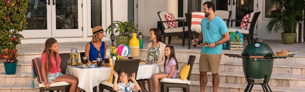 A family enjoys a sunny backyard gathering by the pool. Two women and two children sit around an outdoor dining table set with drinks and snacks, while a man grills food on a Big Green Egg. The patio is decorated with potted plants, bright cushions, and summer décor, creating a relaxed and cheerful atmosphere.
