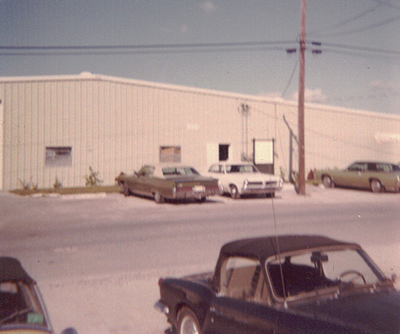 Vintage photo of the original Pinch A Penny warehouse with classic cars parked out front on a sunny day.
