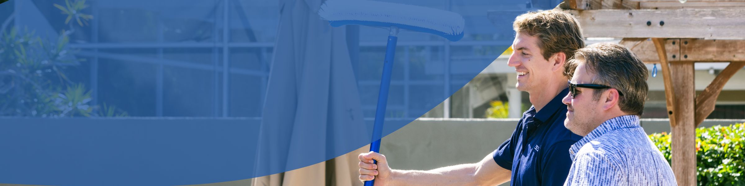 Two men standing near swimming pool, one holding a long pool brush pole