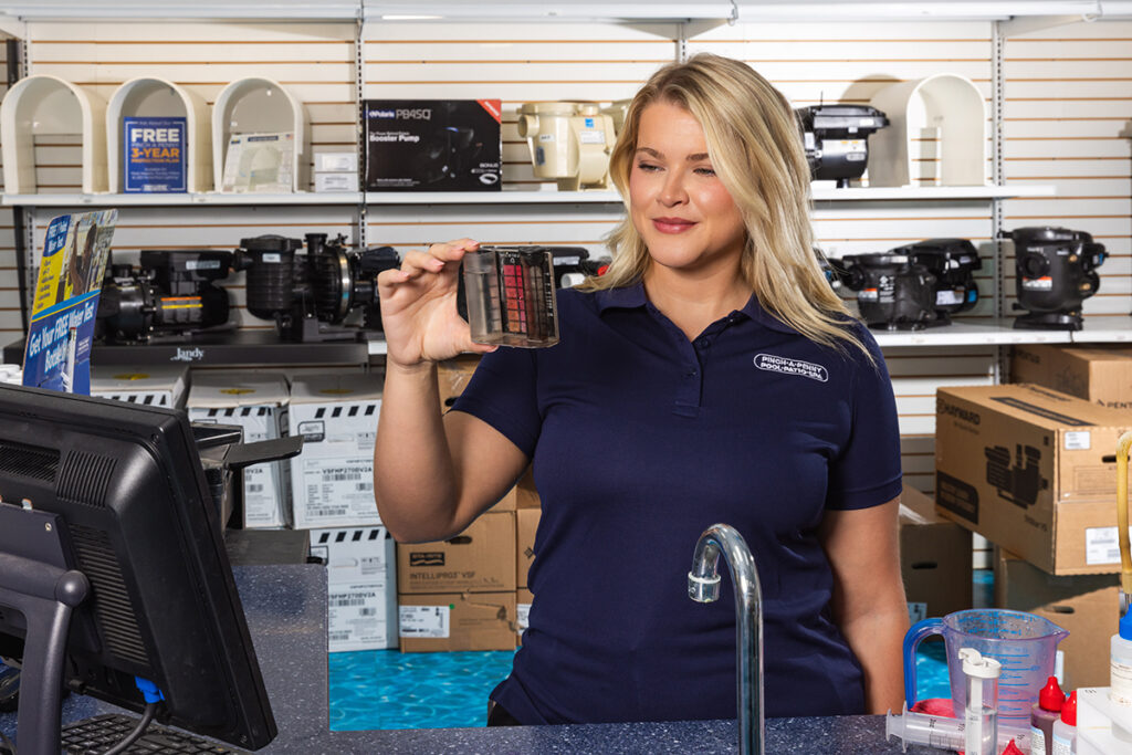 Pinch A Penny team member in a navy polo shirt testing pool water with a color comparison kit at a store counter, with pool equipment displayed on shelves behind her.