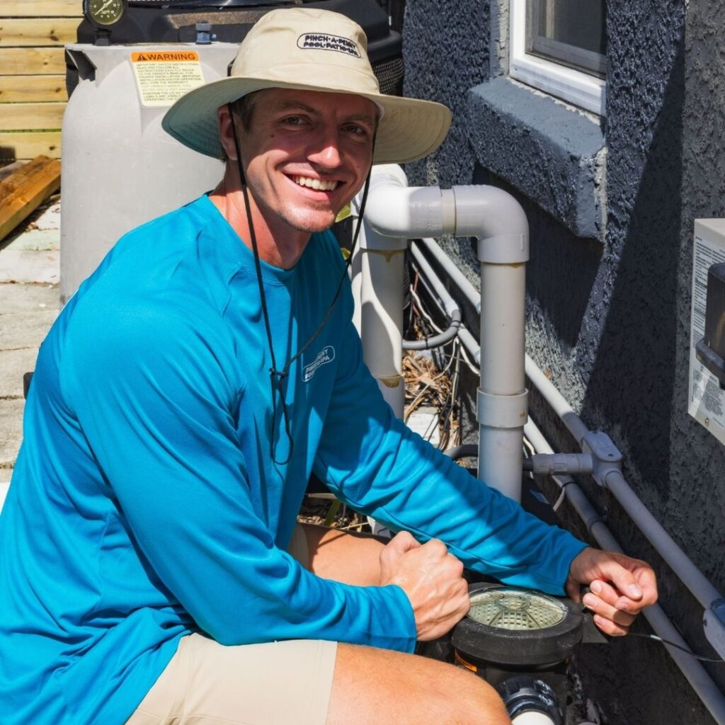 Smiling Pinch A Penny team member in a wide-brim hat and blue long-sleeve shirt kneeling beside pool equipment and plumbing while performing maintenance outside a home.