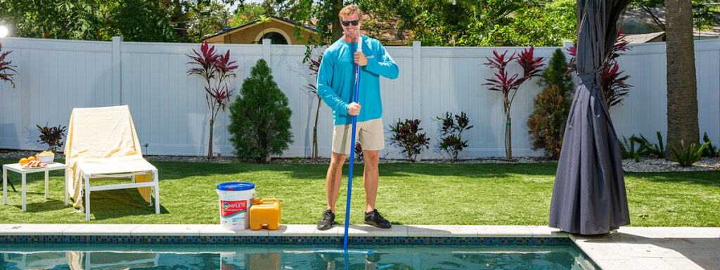 Pinch A Penny team member in a blue long-sleeve shirt skimming leaves from a backyard swimming pool, with pool chemicals and equipment nearby on the patio.