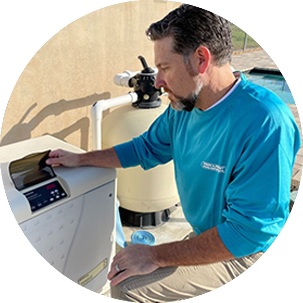 Pinch A Penny technician kneels beside pool equipment, opening the control panel on a pool heat pump near a filter tank at a backyard pool.