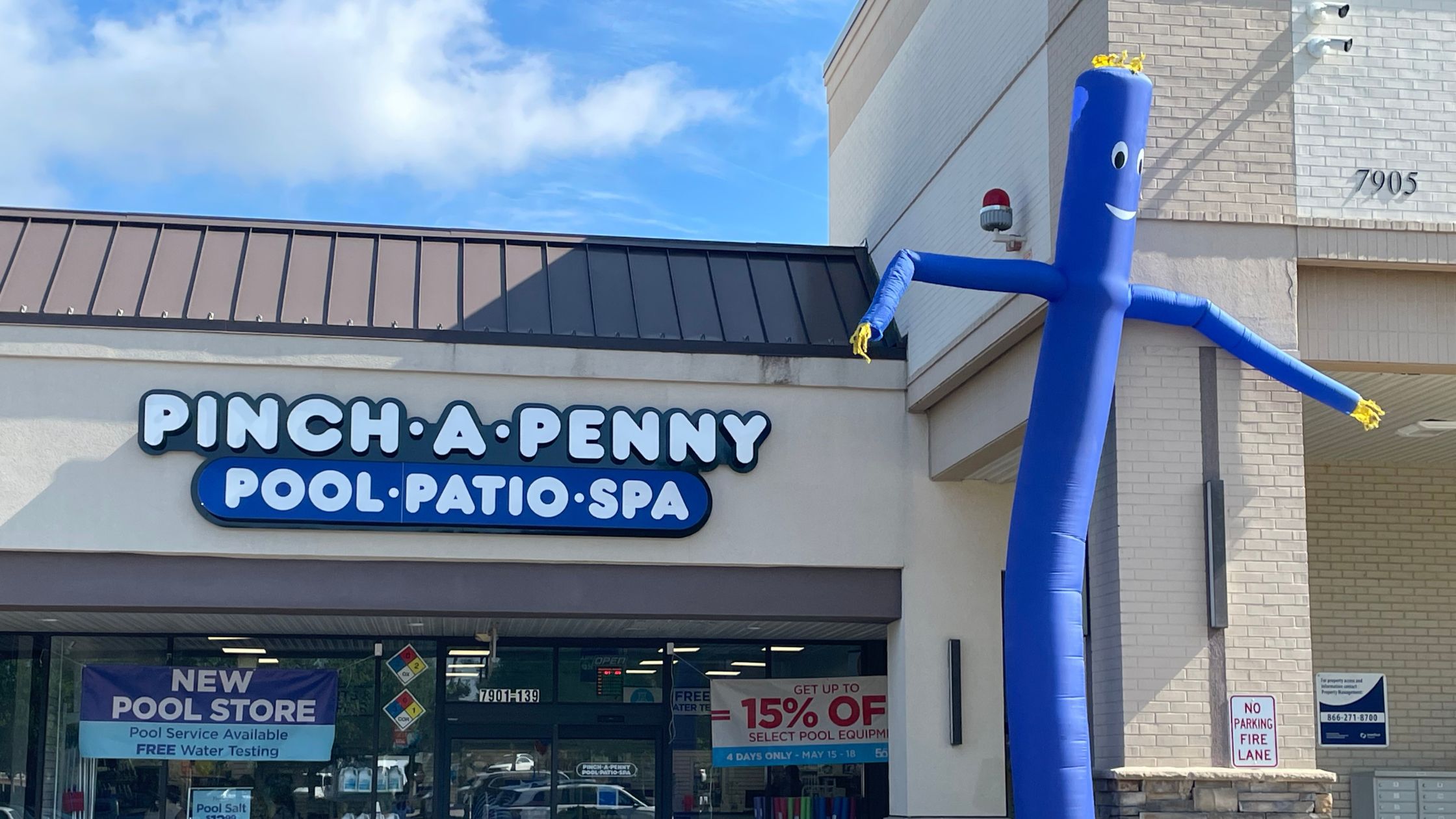 The storefront of a Pinch A Penny Pool Patio Spa retail location. A tall, bright blue inflatable tube man stands to the right of the entrance under a clear blue sky. Banners in the window advertise free water testing and pool equipment discounts.