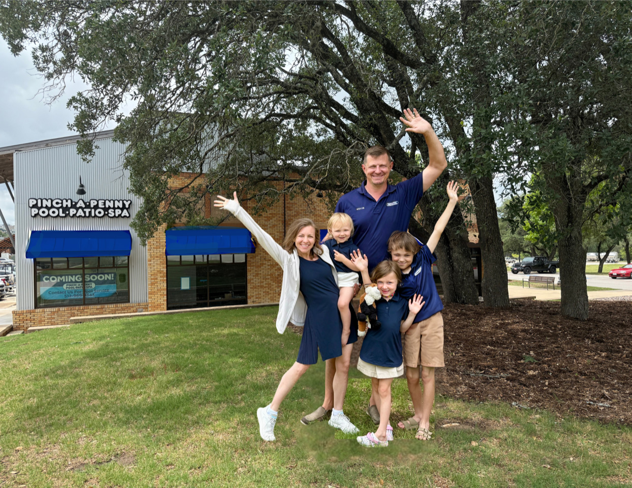 A cheerful family portrait of owners Kevin and Cheryl Bell with their three children waving and posing on a grassy lawn in front of their new store 295, a Pinch A Penny location featuring a "Coming Soon!" sign under large shade trees.