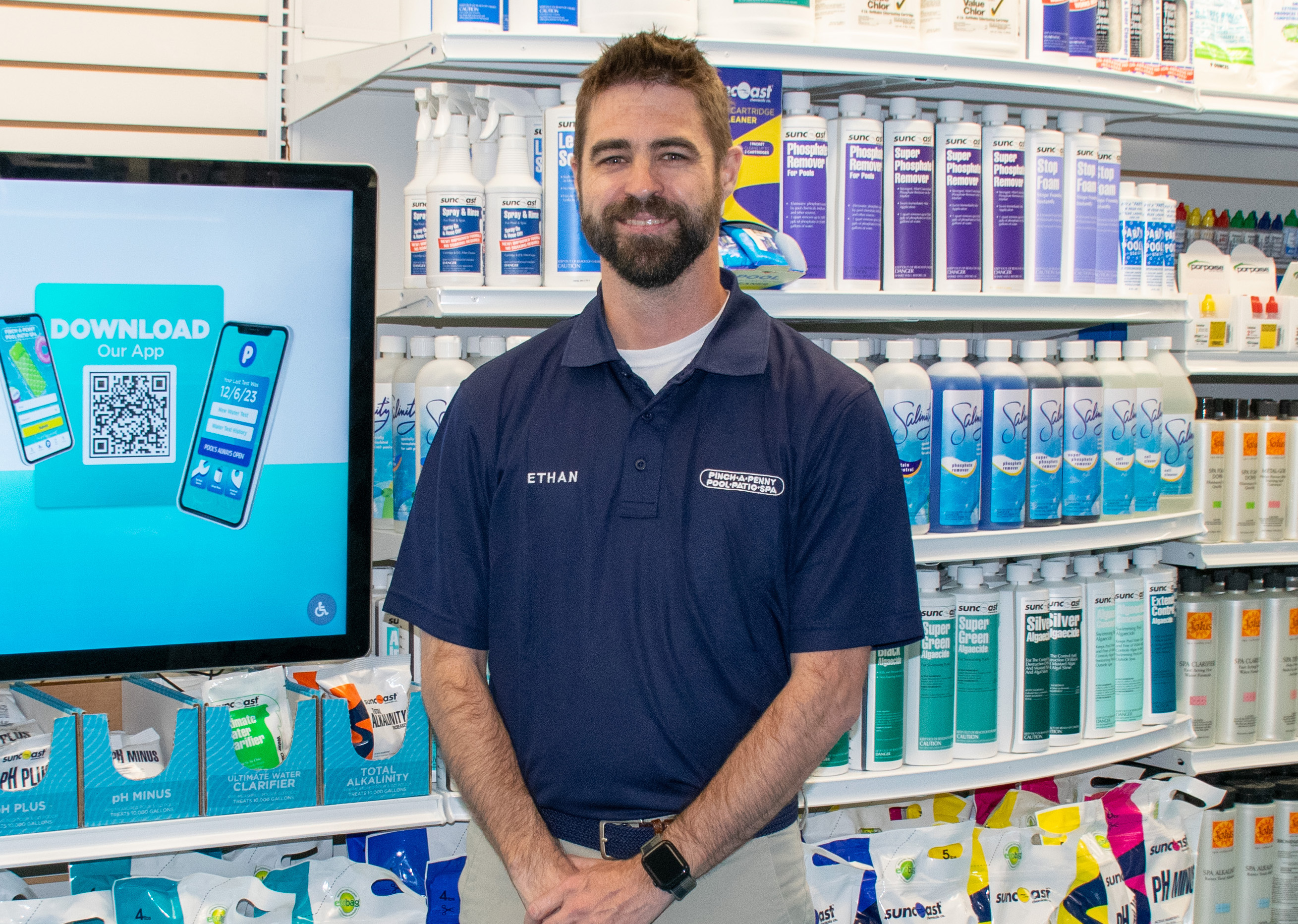 A portrait of a smiling man named Ethan, the owner of store 307, standing inside a Pinch A Penny retail location with his hands clasped in front of him. He wears a navy blue company polo shirt and stands next to a digital display promoting the company app, with fully stocked shelves of pool chemicals behind him.