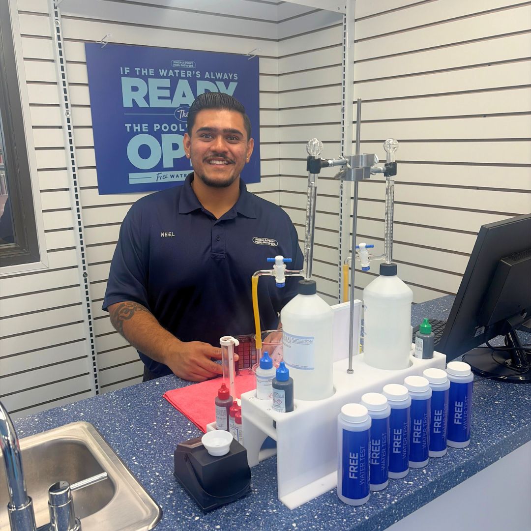 A professional portrait of store 309 owner Neel Patel smiling behind the water testing counter inside his Pinch A Penny retail location. He wears a navy blue company polo shirt embroidered with his name, posing next to professional water analysis equipment, bottles labeled "FREE WATER TEST", and a blue wall sign reading "If the water's always ready, the pool's always open."