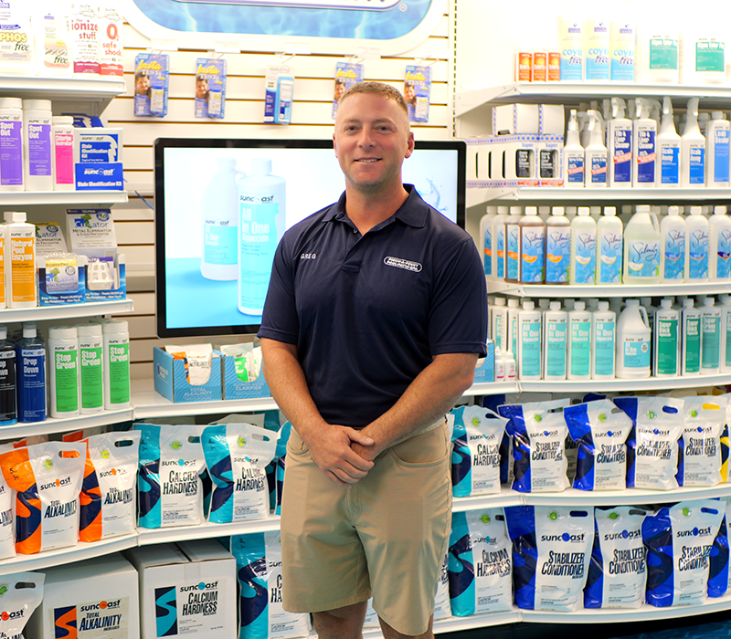 A professional portrait of store 312 owner Greg standing with his hands clasped inside his Pinch A Penny retail store. He wears a navy blue company polo shirt embroidered with his name and stands against a backdrop of fully stocked shelves filled with Suncoast pool chemicals and a digital display screen.