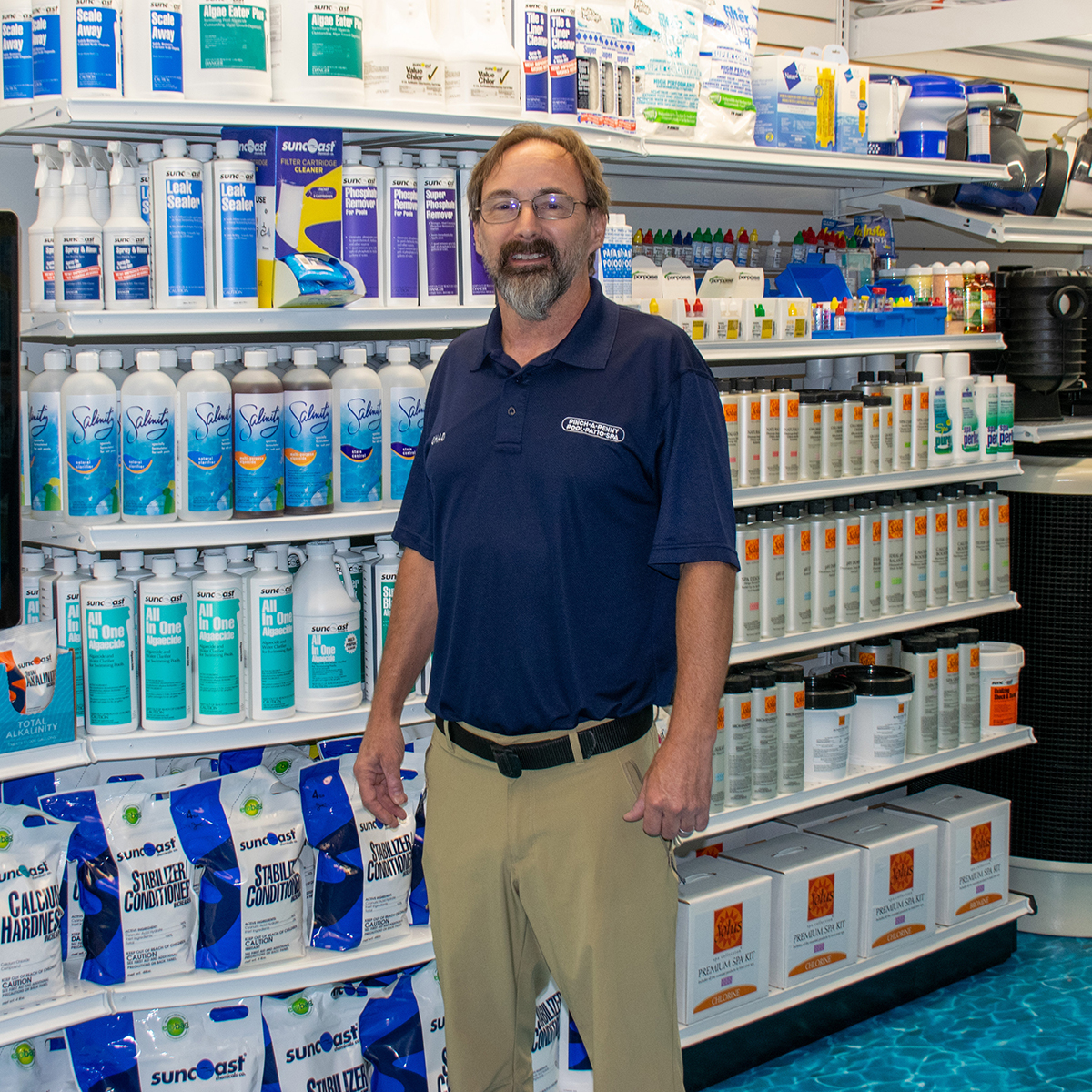 A professional portrait of store 313 owner Chad standing inside his Pinch A Penny retail location. He wears glasses, a goatee, and a navy blue company polo shirt paired with khaki pants, posing in front of brightly lit retail shelves stocked with Suncoast pool chemicals and maintenance products.